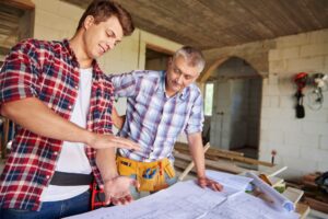 Two men in plaid shirts review construction blueprints on a worktable in a workshop setting.