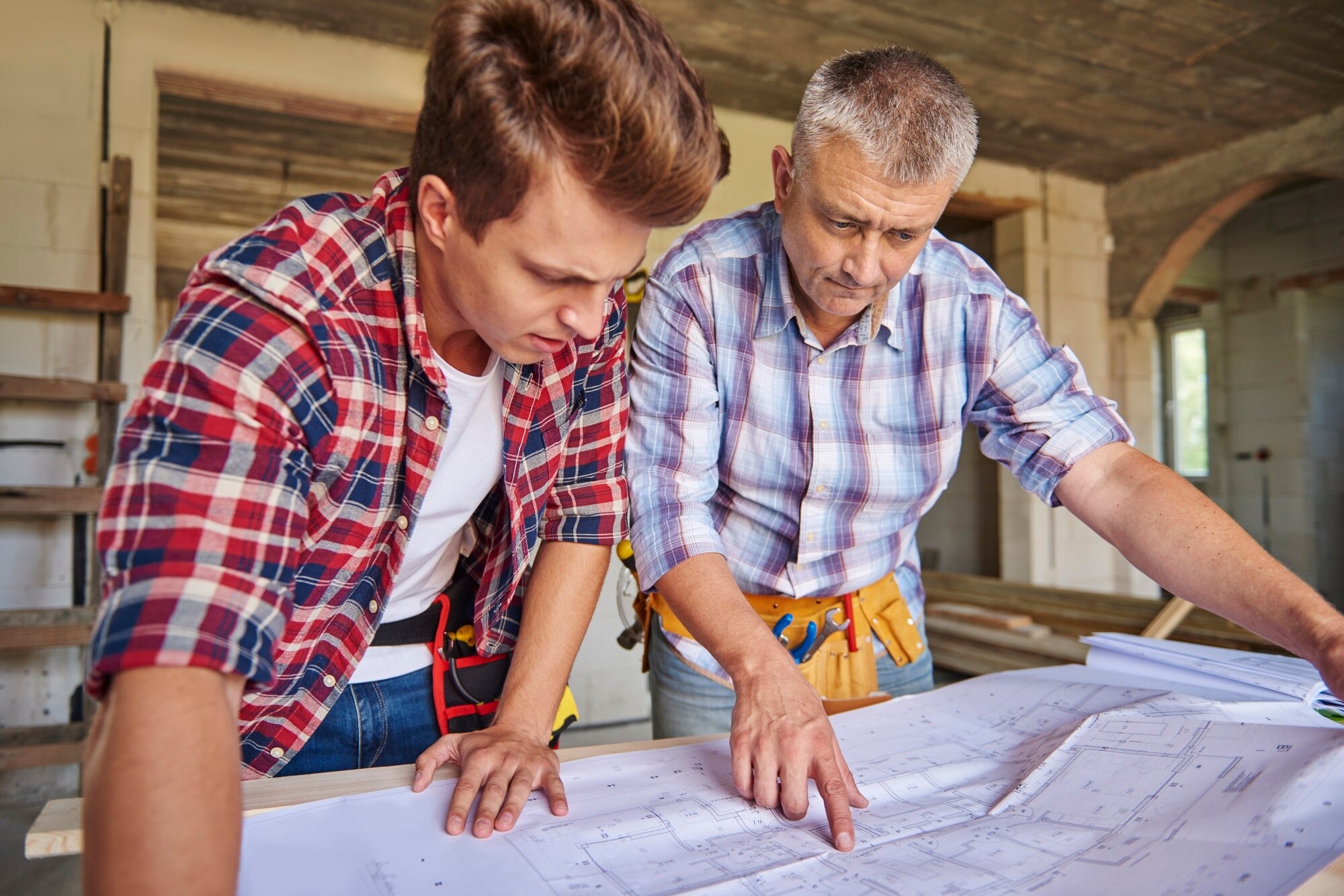Two men in plaid shirts study blueprints on a table in a building under construction.