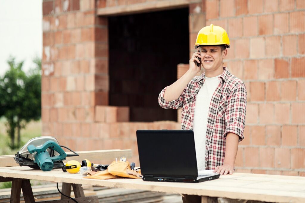 Construction worker in a yellow hard hat talking on a mobile phone beside a laptop on a workbench with tools nearby at a building site. (informative)